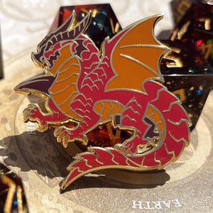 K&ouml;nnte beinhalten: Ein roter, orangefarbener und goldener Drachen-Emaille-Pin mit goldenem Umriss. Der Drache befindet sich in einer dynamischen Pose mit ausgebreiteten Fl&uuml;geln. Der Pin befindet sich vor einem Hintergrund aus W&uuml;rfeln.