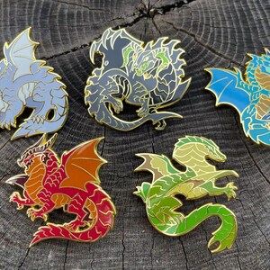 K&ouml;nnte beinhalten: F&uuml;nf farbenfrohe Drachen-Emaille-Anstecknadeln mit goldfarbenen Umrissen. Die Drachen sind in Blau-, Grau-, Rot-, Orange- und Gr&uuml;nt&ouml;nen gehalten. Die Anstecknadeln werden auf einer strukturierten Holzoberfl&auml;che pr&auml;sentiert.