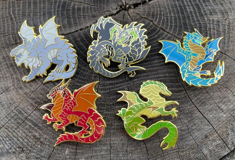 K&ouml;nnte beinhalten: F&uuml;nf farbenfrohe Drachen-Emaille-Anstecknadeln mit goldfarbenen Umrissen. Die Drachen sind in Blau-, Grau-, Rot-, Orange- und Gr&uuml;nt&ouml;nen gehalten. Die Anstecknadeln werden auf einer strukturierten Holzoberfl&auml;che pr&auml;sentiert.