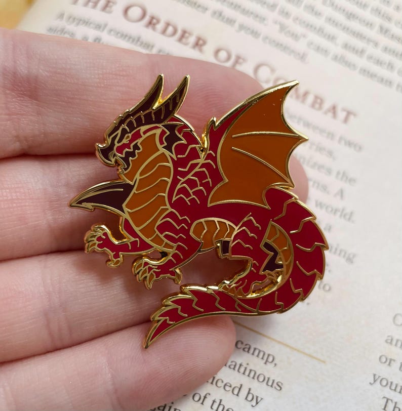 K&ouml;nnte beinhalten: Ein detaillierter Emaille-Pin mit einem roten und goldenen Drachen mit ausgebreiteten Fl&uuml;geln. Der Drache hat aufwendige Schuppen und einen goldenen Umriss. Der Pin ist ein Accessoire mit Fantasy-Thema, perfekt f&uuml;r Jacken, Taschen oder Sammlungen.