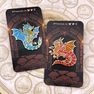 K&ouml;nnte beinhalten: Zwei Emaille-Anstecknadeln mit Drachendesign. Ein Drache ist blau und gold, der andere rot und gold. Jede Anstecknadel befindet sich auf einer Karte mit dem Text "@Toadstown on" und einem Fantasy-Kartenhintergrund.