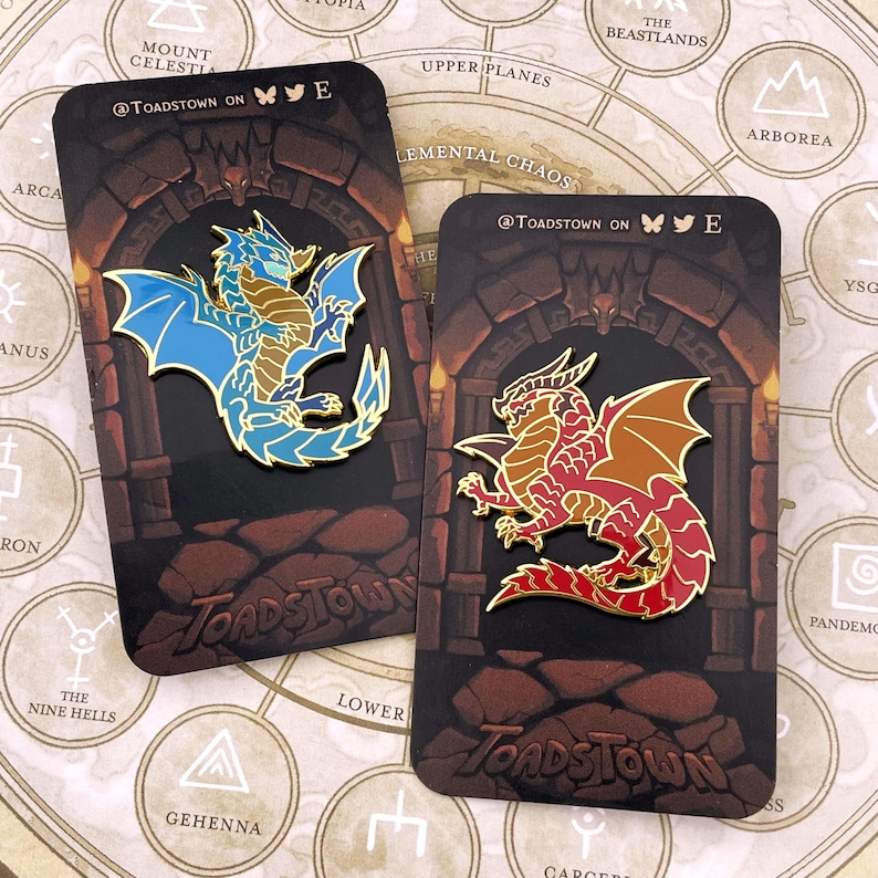 K&ouml;nnte beinhalten: Zwei Emaille-Anstecknadeln mit Drachendesign. Ein Drache ist blau und gold, der andere rot und gold. Jede Anstecknadel befindet sich auf einer Karte mit dem Text "@Toadstown on" und einem Fantasy-Kartenhintergrund.