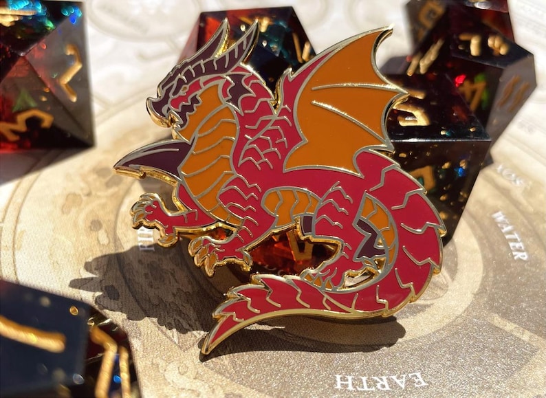 K&ouml;nnte beinhalten: Ein roter, orangefarbener und goldener Drachen-Emaille-Pin mit goldenem Umriss. Der Drache befindet sich in einer dynamischen Pose mit ausgebreiteten Fl&uuml;geln. Der Pin befindet sich vor einem Hintergrund aus W&uuml;rfeln.