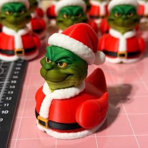 Könnte beinhalten: Eine grüne Quietscheente im Grinch-Design, die einen roten Weihnachtsmannanzug mit weißer Bordüre und einem schwarzen Gürtel mit goldener Schnalle trägt. Die Ente hat einen roten Weihnachtsmannhut mit weißer Bordüre. Weitere ähnliche Enten sind im Hintergrund.