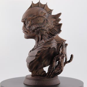 May include: A detailed bust of a creature with a fish-like head and textured skin, in a brown finish. The sculpture features intricate details, including fins and a hand-like appendage. The bust is mounted on a circular base.