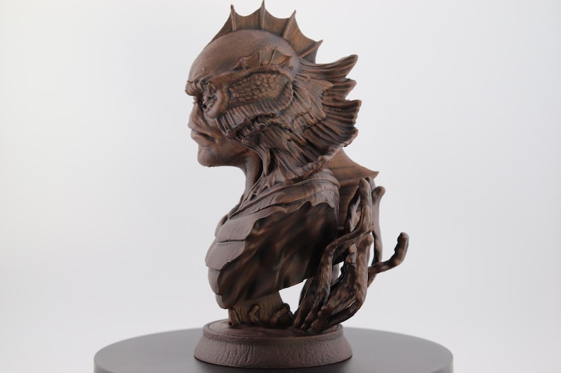May include: A detailed bust of a creature with a fish-like head and textured skin, in a brown finish. The sculpture features intricate details, including fins and a hand-like appendage. The bust is mounted on a circular base.