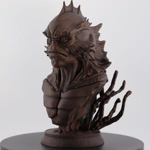 May include: A detailed brown bust of a creature with a fish-like head, spiky fins, and a textured body. The sculpture has a stern expression and is mounted on a round base with stylized aquatic elements.