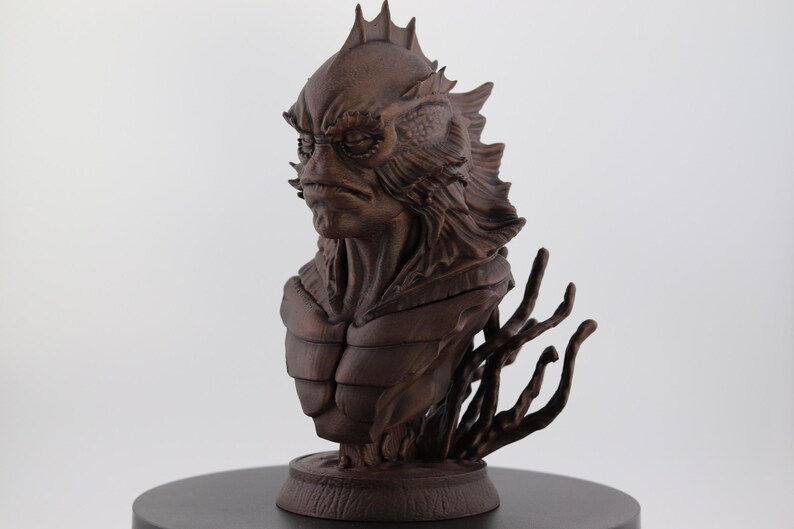 May include: A detailed brown bust of a creature with a fish-like head, spiky fins, and a textured body. The sculpture has a stern expression and is mounted on a round base with stylized aquatic elements.