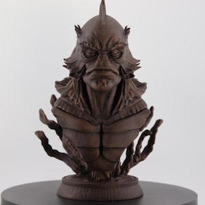 May include: A detailed brown bust of a creature with a fish-like head, prominent brow ridges, and textured skin. The sculpture features intricate details and is mounted on a round base. The creature has a fin on top of its head and is surrounded by aquatic-like appendages.