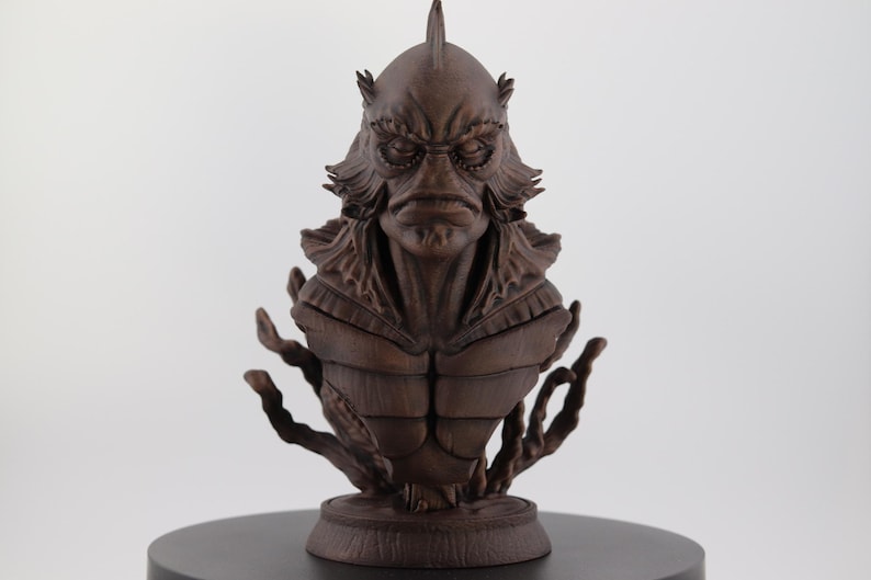 May include: A detailed brown bust of a creature with a fish-like head, prominent brow ridges, and textured skin. The sculpture features intricate details and is mounted on a round base. The creature has a fin on top of its head and is surrounded by aquatic-like appendages.