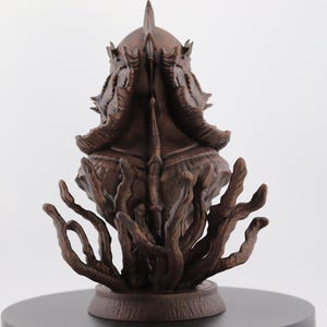 May include: A detailed brown sculpture of a creature with a crown-like head and multiple tentacles. The sculpture is set on a dark circular base against a white background. The intricate design suggests a fantasy or mythological theme.