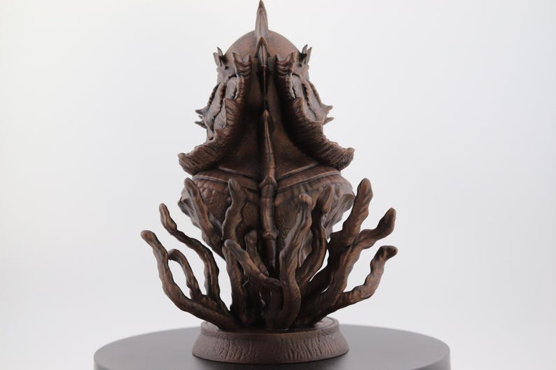 May include: A detailed brown sculpture of a creature with a crown-like head and multiple tentacles. The sculpture is set on a dark circular base against a white background. The intricate design suggests a fantasy or mythological theme.