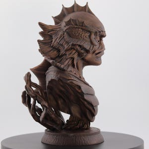 May include: A detailed, brown sculpture of a creature's bust, featuring intricate details of the head, face, and shoulders. The sculpture is mounted on a round, dark base, showcasing the creature's profile with textured elements.
