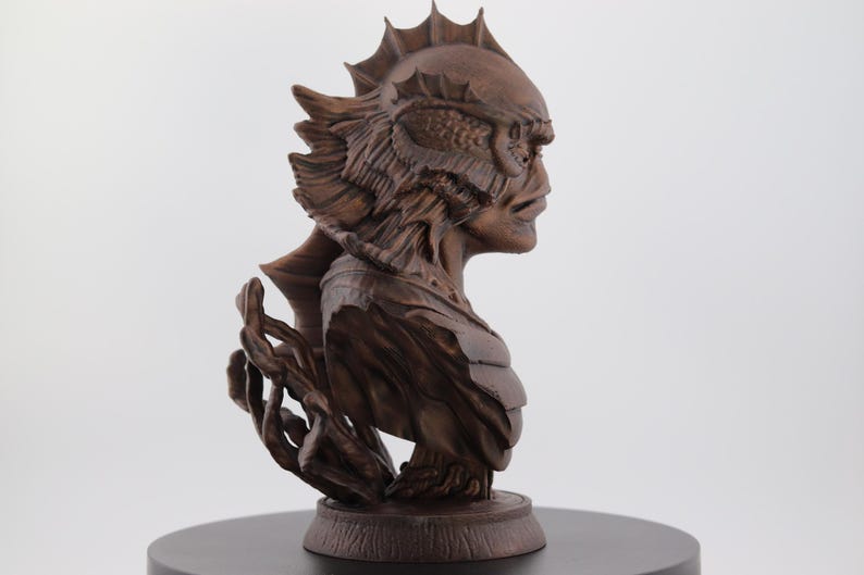 May include: A detailed, brown sculpture of a creature's bust, featuring intricate details of the head, face, and shoulders. The sculpture is mounted on a round, dark base, showcasing the creature's profile with textured elements.