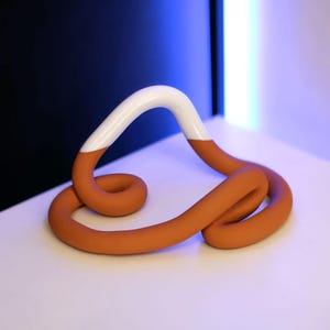 Könnte beinhalten: Eine terracottafarbene Keramikskulptur mit einer weißen Glasur auf der oberen Hälfte. Die Skulptur ist wie eine durchgehende Schleife mit einer Drehung in der Mitte geformt.