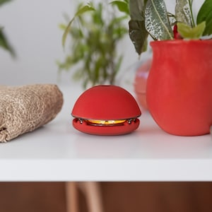 May include: A red, round, electric candle with a warm, yellow glow. The candle is on a white table with a brown woven bag and potted plants in the background.