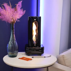 Puede incluir: Una chimenea de mesa negra con una ventana de vidrio que muestra una llama en su interior. La chimenea está sobre una mesa blanca con un jarrón de flores rosas y un pequeño objeto plateado.
