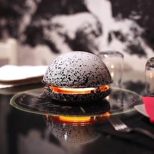 Könnte beinhalten: Ein gesprenkelter, kugelförmiger Kerzenhalter mit orangefarbenem Schein. Der Halter steht auf einer reflektierenden Oberfläche, mit einer Gabel, einem Teller und einem Glas im Hintergrund. Das Gesamtthema ist Essen und Ambiente. Der Kerzenhalter hat einen Durchmesser von etwa 10 cm.