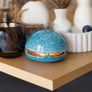 Könnte beinhalten: Ein tealblauer Keramikdiffusor mit silbernem Rand und einem warmen orangenen Schein, der von unten ausstrahlt. Der Diffusor steht auf einer hellbraunen Holzoberfläche.