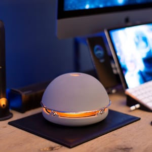 Könnte beinhalten: Ein grauer, kuppelförmiger Diffusor mit einem silbernen Metallband um die Mitte. Der Diffusor leuchtet in einem warmen Orangeton.