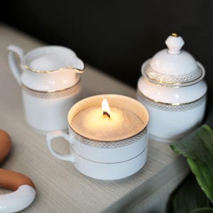 Könnte beinhalten: Eine weiße Teetassenkerze mit goldener Verzierung und einem brennenden Docht. Die Kerze steht auf einem Holztisch neben einem weißen Milchkännchen und einer weißen Zuckerdose mit Deckel.