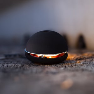 Könnte beinhalten: Ein schwarzer Keramikdiffusor mit einem silbernen Metallband um die Mitte. Der Diffusor ist beleuchtet und leuchtet orange.