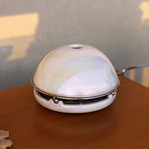 Puede incluir: Un difusor de cerámica blanca con un borde de metal plateado y un acabado iridiscente. El difusor está sentado en una mesa de madera.