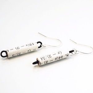 Puede incluir: Un par de pendientes de plata con ganchos negros. Cada pendiente presenta un trozo de papel enrollado con ecuaciones matemáticas impresas en él. Las ecuaciones son: 12(2-2) = (2-3)/3 y 8. 54(x - 3) + (x - 3)/54.