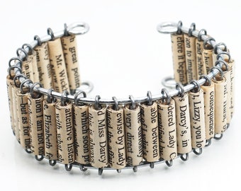 May include: A silver wire bracelet with rolled up paper beads. Each bead has text from a book printed on it. The text includes "Lizzy, you lo", "Darcy's, in", "consequence", "able between", "very direct qu", "ewise by Lady", "lated the affair", "d Miss Darcy", "this is always", "which soon pr", "inst another", "attentive listen", "Mr. Wickh", "Every did with what sh", "jom who", "in in", "e. Elizabeth fe", "ion I am not on", "but as for the", "Catherine za".