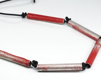 May include: A necklace featuring alternating red and silver-colored cylindrical beads. The beads are connected by black cord, creating a unique and modern design. The necklace is displayed against a white background.