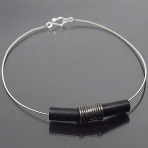 May include: A minimalist necklace with a thin, silver-coloured wire. The necklace has a black cylindrical centrepiece with silver-coloured metal rings and a hook clasp. The jewellery is displayed on a reflective surface.