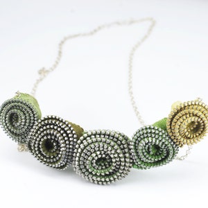 Puede incluir: Un collar verde y dorado hecho de cremalleras enrolladas. El collar tiene una cadena plateada y está hecho de materiales reciclados.