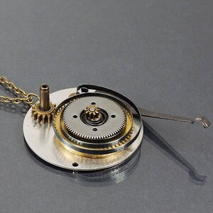 May include: A silver and gold steampunk necklace with a chain and a pendant featuring a circular gear mechanism with a small spring.