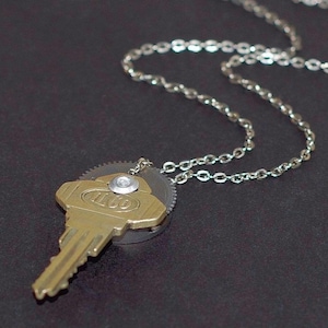 Puede incluir: Un collar único con un colgante de llave de latón con la palabra "ILCO" y una cadena de plata. La llave está unida a un pequeño engranaje y asegurada con un remache plateado. La cadena es de eslabones de plata delicados.