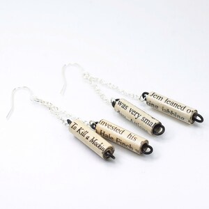 May include: A pair of silver chain earrings with three rolled paper charms. The charms are made from book pages and feature the text "Jem leaned o", "king iahhing", "was very smal", "day Anicu", "invested his", "Hale Finch w", and "To Kill a Mockin".