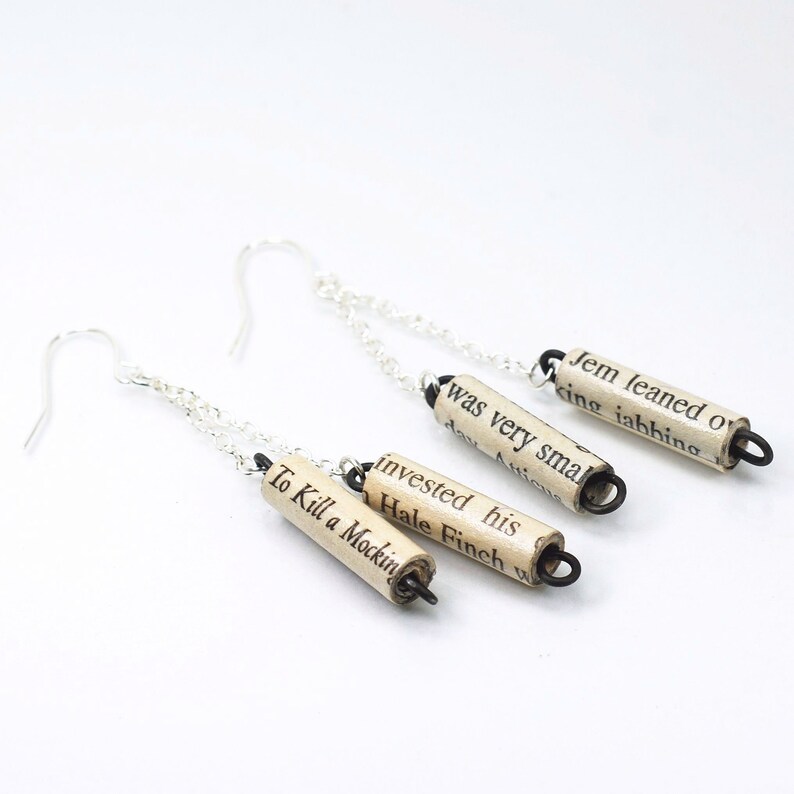 May include: A pair of silver chain earrings with three rolled paper charms. The charms are made from book pages and feature the text "Jem leaned o", "king iahhing", "was very smal", "day Anicu", "invested his", "Hale Finch w", and "To Kill a Mockin".