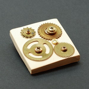 Puede incluir: Primer plano de una baldosa rectangular blanca con cinco engranajes de latón de diferentes tamaños y formas. Los engranajes están dispuestos en un patrón decorativo y tienen un aspecto desgastado y antiguo.