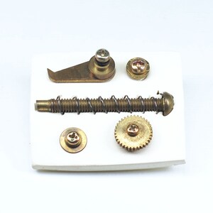 May include: A set of five brass-colored metal parts, including a spring, a gear, a hook, and two screws, displayed on a white background.