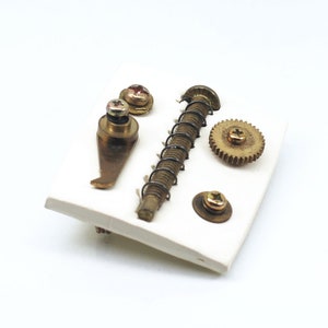May include: A white ceramic tile with a collection of small brass gears, screws, and a spring. The items are arranged in a random pattern.