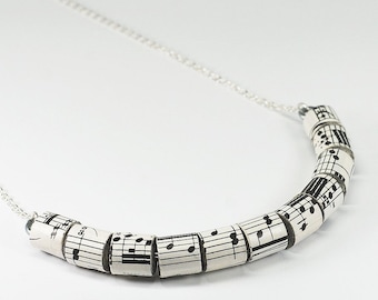 May include: A silver chain necklace with a pendant made of rolled up sheet music. The sheet music is black and white and features musical notes.