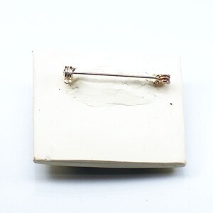 May include: A silver pin with a gold-toned clasp. The pin is on a white surface.