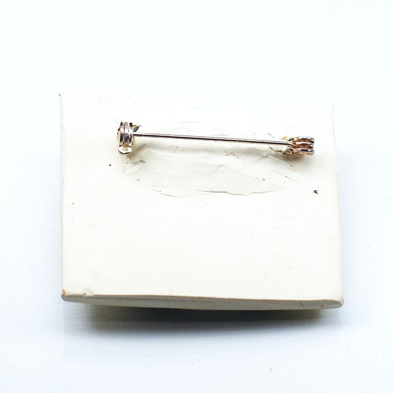 May include: A silver pin with a gold-toned clasp. The pin is on a white surface.