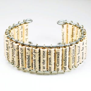 May include: A unique cuff bracelet made from rolled paper strips with printed text, held together by a silver-colored wire frame. The paper has a cream color, and the text is black. The bracelet has an open design.
