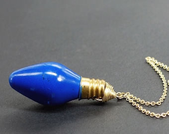 Peut inclure: Un collier avec un pendentif en forme d'ampoule bleue et une chaîne dorée. L'ampoule a la forme d'une ampoule de Noël classique.