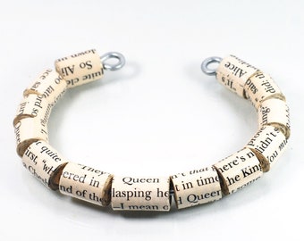 May include: A white bracelet made of rolled-up pages from a book. The bracelet is made of several beads, each with a different word or phrase from the book. The bracelet is closed with two metal clasps.