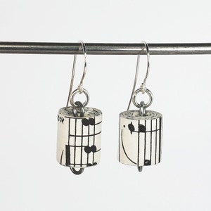 May include: A pair of silver dangle earrings with cylindrical charms made from rolled sheet music. The charms feature black musical notes on a white background.