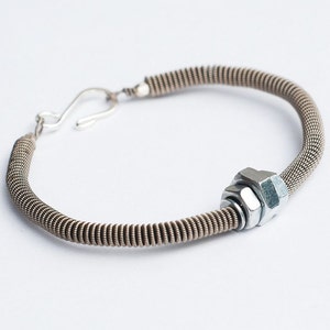 May include: A silver-toned bracelet with a unique industrial design. The bracelet features a coiled wire band with a hex nut and washer accent. A hook clasp secures the bracelet. The bracelet is shown on a white background.
