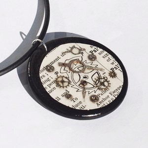 Puede incluir: Un collar con un colgante redondo de madera negra. El colgante contiene un papel blanco con texto y una esfera de reloj de estilo steampunk con engranajes y manecillas. El texto dice "Instrument used by rs; ding; ps e pen nce et ll and To duce iti- a Pal and SIE of wi Pal Pallet Anchor Escape- ant: pp Pall".