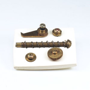 May include: A set of brass hardware parts for a mechanical device, including a small gear, a spring, a screw, a lever, and a small round piece. The parts are arranged on a white surface.