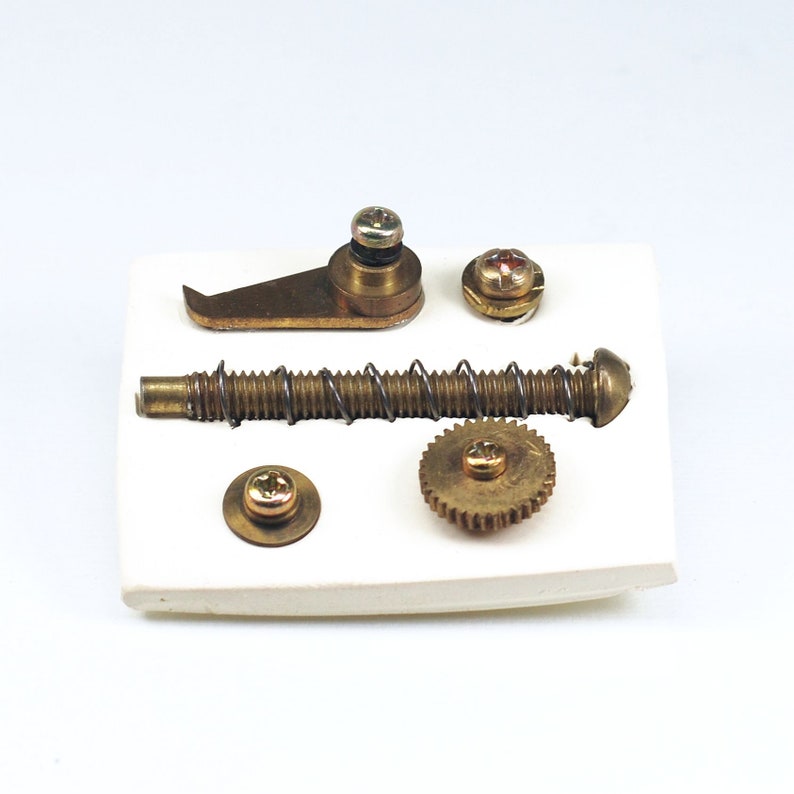 May include: A set of brass hardware parts for a mechanical device, including a small gear, a spring, a screw, a lever, and a small round piece. The parts are arranged on a white surface.